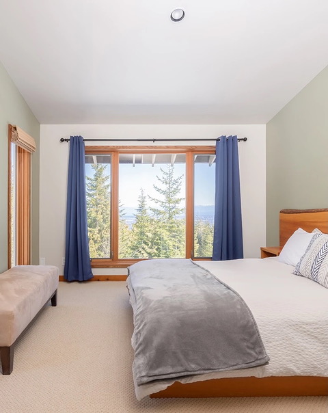 Private bedroom at Halo Ridge Recovery with a made bed, blue curtains, and windows looking through fir trees toward the valley